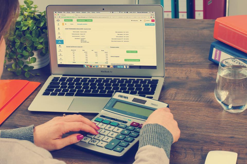 Hands using a calculator next to a laptop showing a spreadsheet of bills